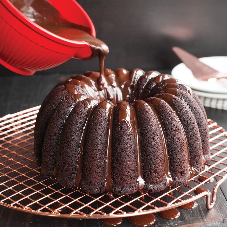 Melted chocolate poured onto a cake from a red pourer. The cake sists on a cooking rack on a wooden surface.