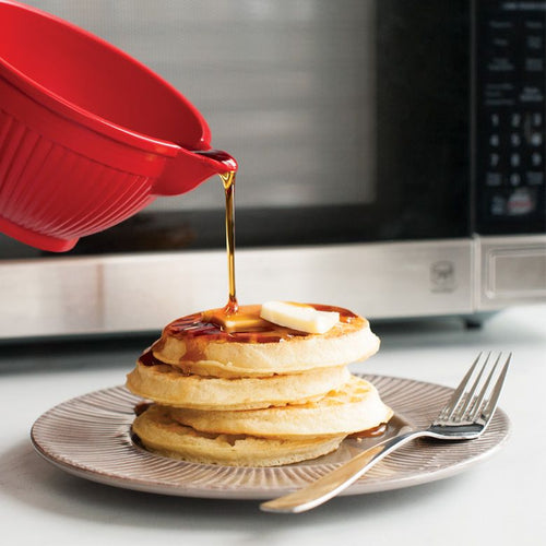 Syrup being poured from a red container onto a stack of waffles on a plate with a fork.