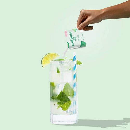 A packet of Craftmix Mint Mojito Cocktail/Mocktail Drink Mixer is being held above a glass filled with ice, lime, and mint leaves, creating a visual representation of how the drink is made.