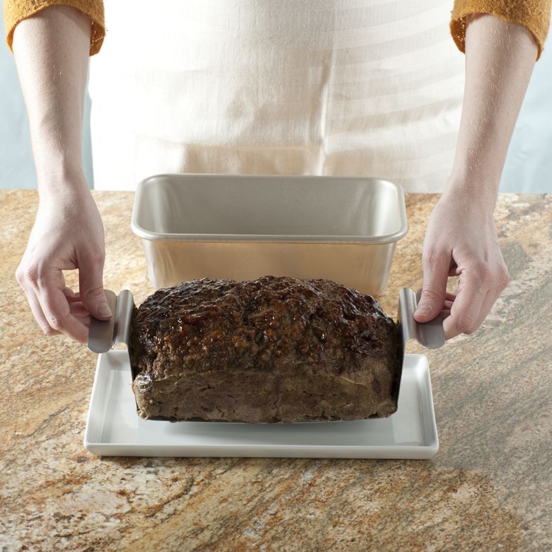 A person using a lifting trivet to put meatloaf on a tray with a meatloaf pan behind it.