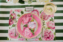 A place setting at a dining table featuring a peony patterned placemat, pink plate peony place card, fork, knife, and name card all on a green striped table runner.