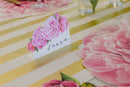 A place card with the name Laura on it with peonies on a gold and white striped table runner.