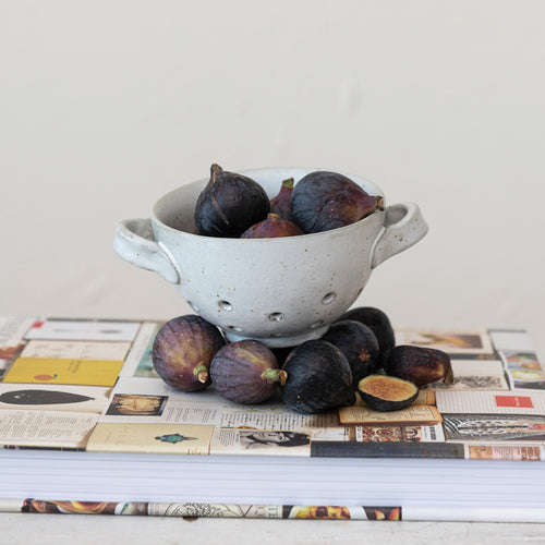 A glazed bowl with handles and holes in the bottom hold figs and sit atop figs on a book.